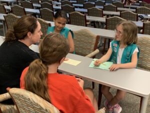 Two Junior Girl Scouts sitting at a table facing adult woman in black sweater and woman with a ponytail in a red shirt.